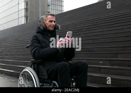 Konzept der behinderten Person. Mann im Rollstuhl draußen auf der Straße vor der Treppe. Menschen, die Technologie mit dem Smartphone nutzen. Stockfoto