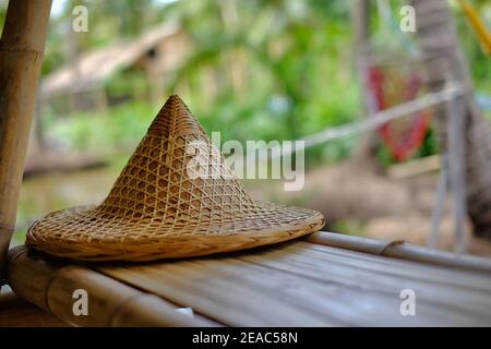 Ein traditioneller südostasiatischer Bauernhut aus gewebtem Bambus und Stroh wird am Rand eines Bambustisches in einer Hütte bei einem Reisfeld in Thailand platziert. Stockfoto