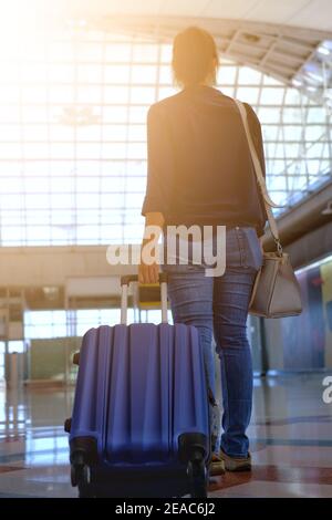 Die Rückansicht einer Frau, die ein Gepäck rollt und zu einem Tor an einem Terminal am Flughafen mit heller Sonne am Gebäudeglas geht. Stockfoto