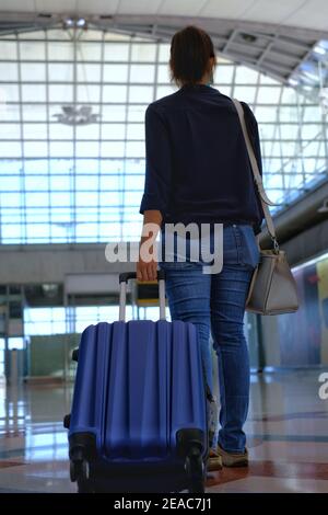 Die Rückansicht einer Frau, die ein Gepäck rollt und zu einem Tor an einem Terminal am Flughafen mit heller Sonne am Gebäudeglas geht. Stockfoto