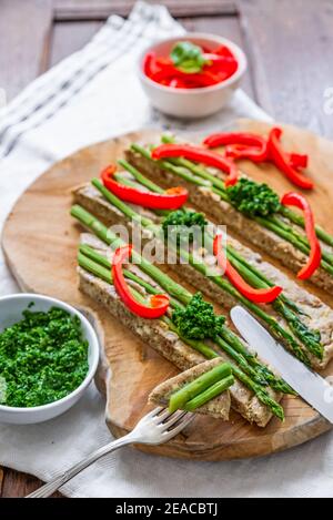 Foccacia mit grünem Spargel, Paprika und grünem Pesto auf einem Holzschneidebrett, Zutaten in kleinen Schüsseln Stockfoto