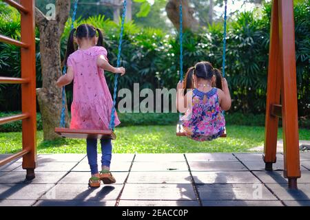 Die Rückansicht zweier junger asiatischer Mädchen, die gemeinsam auf einem Spielplatz spielen, Schaukel reiten und an einem sonnigen Tag gemeinsam ihre Zeit genießen. Stockfoto