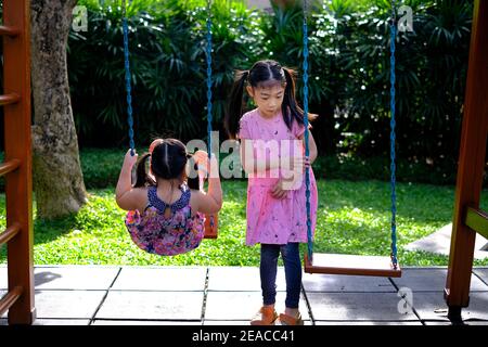 Die Rückansicht zweier junger asiatischer Mädchen, die gemeinsam auf einem Spielplatz spielen, Schaukel reiten und an einem sonnigen Tag gemeinsam ihre Zeit genießen. Stockfoto