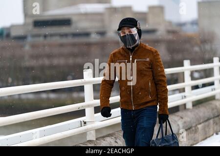 LONDON, ENGLAND - FEBRUAR, 2021: Junger Mann, der die Kamera mit Gesichtsmaske und Gesichtsschild anschaut, läuft auf der Waterloo Bridge in London während eines s Stockfoto