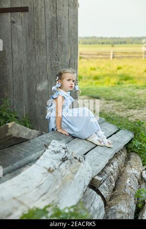 Nettes kleines lustiges Mädchen in einem blauen Kleid sitzt im Hof des Dorfes. Frohe Kindheit. Stockfoto