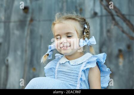 Nettes kleines lustiges Mädchen in einem blauen Kleid sitzt im Hof des Dorfes. Frohe Kindheit. Stockfoto