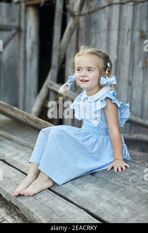 Nettes kleines lustiges Mädchen in einem blauen Kleid sitzt im Hof des Dorfes. Frohe Kindheit. Stockfoto