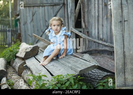 Nettes kleines lustiges Mädchen in einem blauen Kleid sitzt im Hof des Dorfes. Frohe Kindheit. Stockfoto