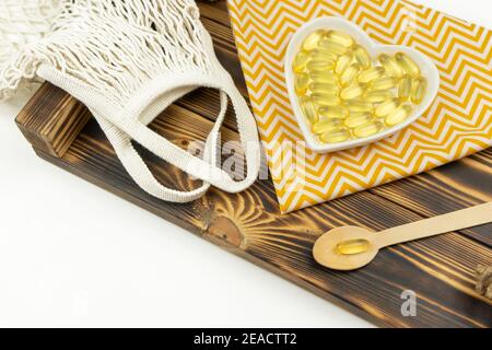Gelbe Kapseln von Omega 3 liegen auf weißer Keramikplatte In Form des Herzens auf einem Holztablett Stockfoto