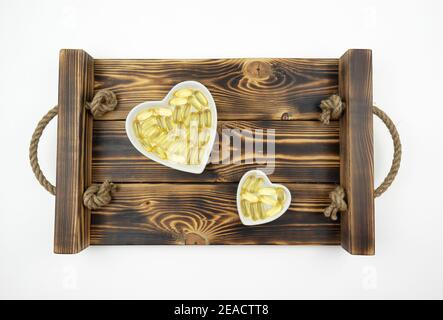 Gelbe Kapseln von Omega 3 liegen auf weißen Keramikplatten In Form der Herzen auf einem Holztablett Stockfoto