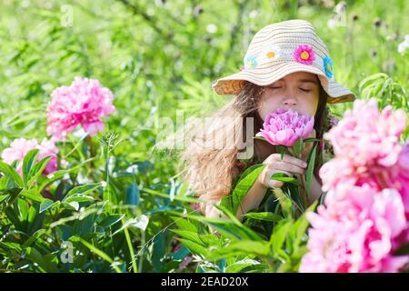 Schönes kleines Mädchen hält eine rosa Pfingstrose an einem sonnigen Tag im Garten. Hübsches Mädchen trägt ein modisch gemustertes Sommerkleid und Strohhut Stockfoto