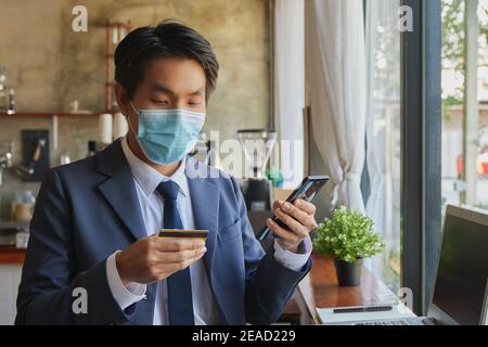 Zoom View Front Right Standing Asian Geschäftsmann in Anzug tragen Gesichtsmaske Verwenden Sie Smartphone für Online-Shopping. Asiatische Geschäftsmann arbeiten von überall mit Stockfoto