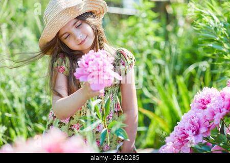 Schönes kleines Mädchen hält eine rosa Pfingstrose an einem sonnigen Tag im Garten. Hübsches Mädchen trägt ein modisch gemustertes Sommerkleid und Strohhut Stockfoto