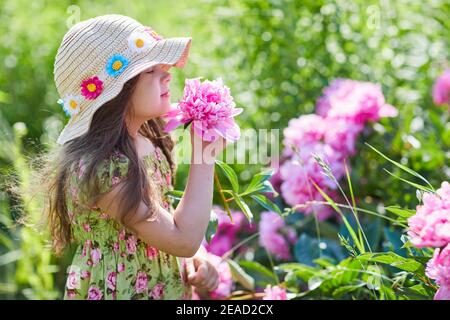 Schönes kleines Mädchen hält eine rosa Pfingstrose an einem sonnigen Tag im Garten. Hübsches Mädchen trägt ein modisch gemustertes Sommerkleid und Strohhut Stockfoto