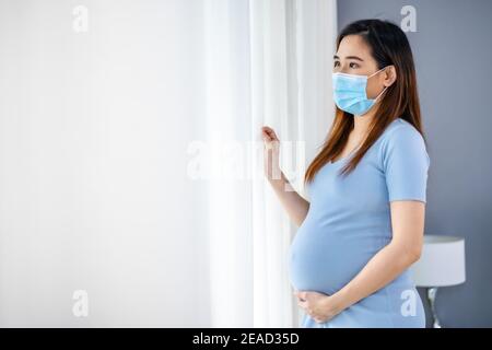 Schwangere Frau in medizinischer Maske und Blick durch Fenster in einem Schlafzimmer, bleiben Sie allein für Schutz Coronavirus (covid-19) Pandemie Stockfoto