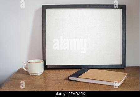 Schwarzer Rahmen Nachbau, auf alten Tisch und weiße Wand, mit leerem Skizzenbuch oder Tagebuch und Kaffeetasse. Auf Holztisch. Stockfoto