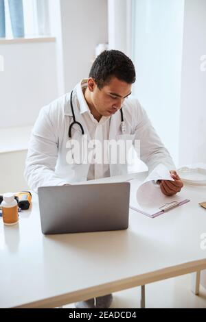 Hart arbeitend ernsthaft Mann Arzt in weißem Gewand, der mit arbeitet Zwischenablage und Laptop im Zimmer im Inneren des medizinischen Zentrums Stockfoto