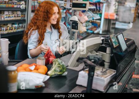 Junge Verkäuferin sitzt an der Kasse im Supermarkt und bedient Käufer. Frau Kassiererin Scannen Lebensmittel an der Kasse. Stockfoto