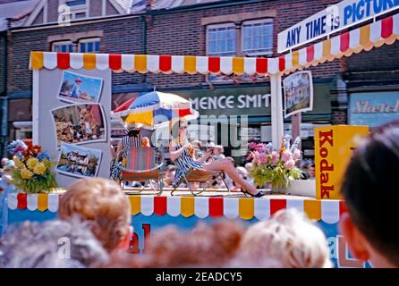 Ein Festwagen für Kodak Film, Verarbeitung und Drucke bei der jährlichen Pfingstparade, Watford, Hertfordshire, England, Großbritannien 1965. Eine Frau in einem Minikleid, die auf einem Strandstuhl sitzt, hält eine Kodak Kamera. Eine riesige Kiste Kodak-Film ist auch auf dem Festwagen. Dieses Bild ist von einem alten Amateur Kodak 35mm Farbtransparenz. Stockfoto