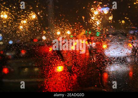 Mehrfarbige Lichter und Regentropfen auf Glas in der abendlichen Hauptverkehrszeit eines Großstadtverkehrs. Stockfoto