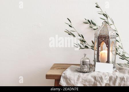 Ramadan Kareem Grußkarte, Einladung Mockup. Silberne Laterne mit brennender Kerze und silberne Tasse mit Tee oder Kaffee. Grüner Olivenbaum verzweigt sich Stockfoto