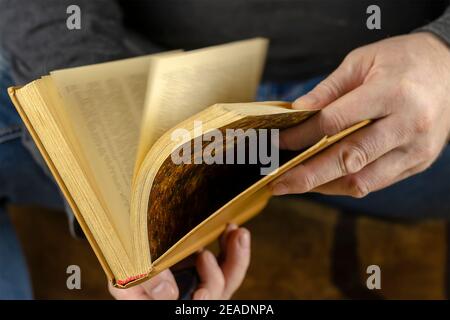 Die Hände eines Mannes blättern durch ein offenes Buch. Ein Hardcover-Buch liegt ihm zu Füßen. Nahaufnahme, selektiver Fokus. Unschärfe in Bewegung Stockfoto