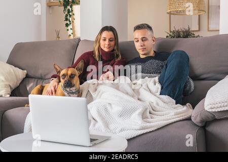 Junges Paar und ihr Hund in gemütlichen Hause, entspannen auf der Couch, während Filme auf Notebook. Lebenskonzept für Paare Stockfoto
