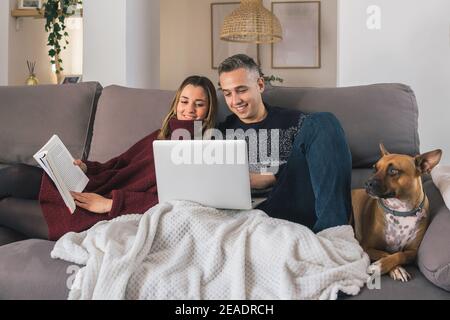 Glückliches Paar und ihr Hund entspannen auf der Couch, ein Buch lesen und mit Laptop, eine tolle Zeit zusammen zu Hause. Lebenskonzept für Paare Stockfoto