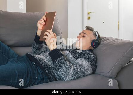 Junger Mann liegt und entspannt auf dem Sofa, während er Musik hört und ein Tablet benutzt. Guy mit tolle Zeit zu Hause Konzept Stockfoto