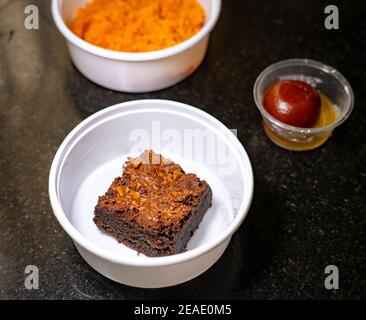 Brownie aus Schokoladenmond in einer weißen Takeout-Box auf schwarzem Granit-Hintergrund. Gulab jamun und Carrot Halwa verschwommen im Hintergrund. Stockfoto
