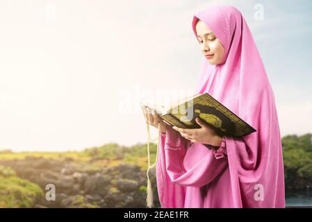 Asiatische muslimische Frau in einem Schleier hält Gebetsperlen und Lesen des Quran im Freien Stockfoto