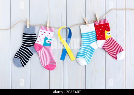 Kindersocken mit Streifen und Farben in Wäscheleine auf Holzhintergrund. In der Mitte befindet sich ein gelbes und blaues Band. World Down Syndrome Day Stockfoto