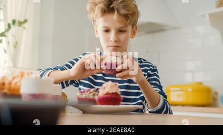 In der Küche: Adorable Boy isst cremigen Cupcake mit Frosting und bestreut Funfetti. Cute Hungry Sweet Tooth Child Bites in Muffin mit zuckerhaltigen Stockfoto