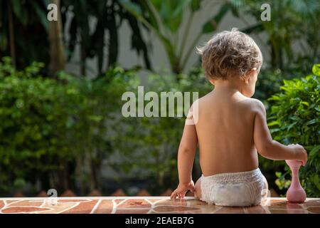 Ein charmantes Kleinkind sitzt mit einem Spielzeug auf den Stufen eines Hauses im Sonnenlicht. Rückansicht. Stockfoto