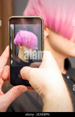 Der Friseur macht Fotos von der kurzen rosa Frisur von Eine junge Frau auf dem Smartphone Stockfoto