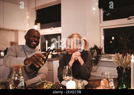 Lachender Mann, der seiner Frau während des Windes ein Glas Wein gießt Ein Candlelight Dinner Party um einen Tisch mit Freunden Stockfoto