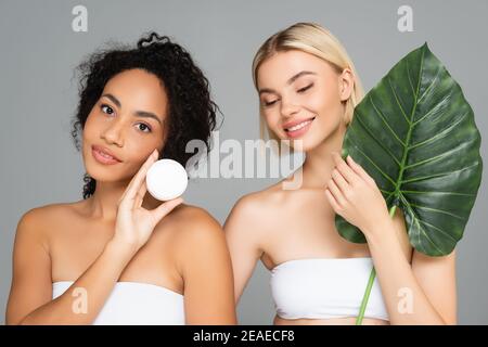 Lächelnde interracial Frauen posiert mit kosmetischen Creme und tropischen Blatt Isoliert auf Grau Stockfoto