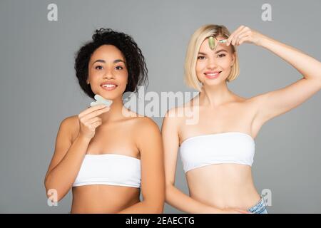 Fröhliche multikulturelle Frauen mit gua Sha und Jade Roller isoliert Auf Grau Stockfoto