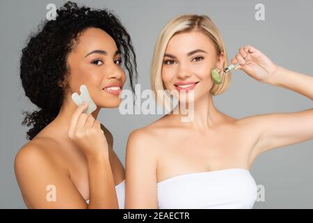 Multiethnische Frauen mit perfekter Haut massieren Gesichter mit Gua Sha Und Jadewalze isoliert auf grau Stockfoto