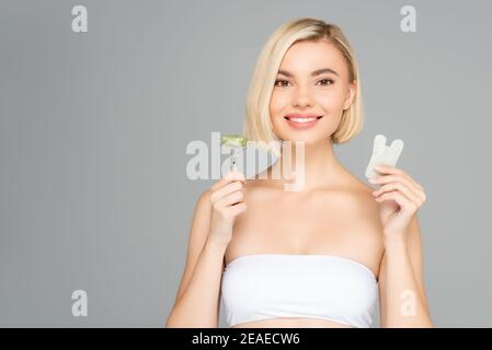 Lächelnde blonde Frau hält Gua Sha und Jade Roller isoliert Auf Grau Stockfoto