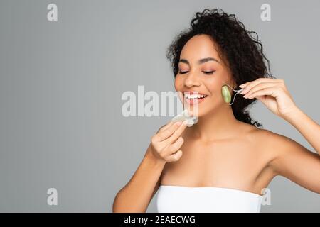 Junge afroamerikanische Frau lächelt, während mit Jade Roller und gua Sha isoliert auf grau Stockfoto