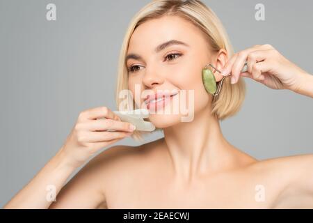 Blonde Frau mit sauberer Haut massieren Gesicht mit Gua Sha Und Jadewalze isoliert auf grau Stockfoto