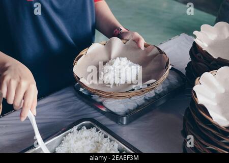 Person Hand legt weißen Reis aus der Stahlpfanne zu Hölzerner Pappteller während des Mittagessens im Restaurant Stockfoto