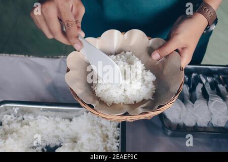 Person Hand legt weißen Reis aus der Stahlpfanne zu Hölzerner Pappteller während des Mittagessens im Restaurant Stockfoto