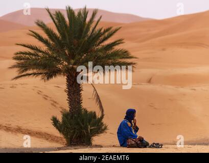 Mann, der unter einer Palme im Sand sitzt sahara Wüste mit blauem Gewand Stockfoto