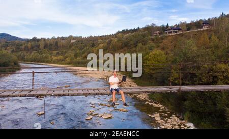 Luftdrohne Ansicht von jungen Mann in weißem Hemd sitzen auf Holzbrücke über Berge Fluss und mit Laptop. Erfolg Freiberufler arbeitet Remote in Bergen, Telearbeit Stockfoto