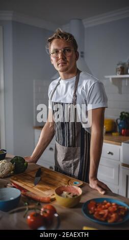 Handsome man in White Shirt und Schürze macht eine gesunde Bio-Salat Mahlzeit in einer modernen sonnigen Küche. Hipster Mann in Brille smiles an der Kamera Stockfoto