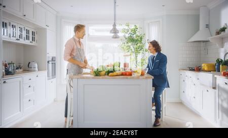 Zwei junge gut aussehende Männer sprechen in der Küche. Einer ist lässig angezogen und sein Freund trägt einen Business-Anzug. Sonnige moderne Küche mit gesundem Grün Stockfoto