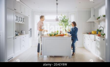 Zwei junge gut aussehende Männer sprechen in der Küche. Einer ist lässig angezogen und sein Freund trägt einen Business-Anzug. Sonnige moderne Küche mit gesundem Grün Stockfoto