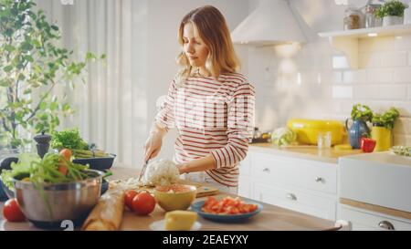 Junge Frau im gestreiften Pullover macht einen gesunden Bio-Salat in einer modernen sonnigen Küche. Authentische Frau hackt einen Blumenkohl mit einem Messer Stockfoto